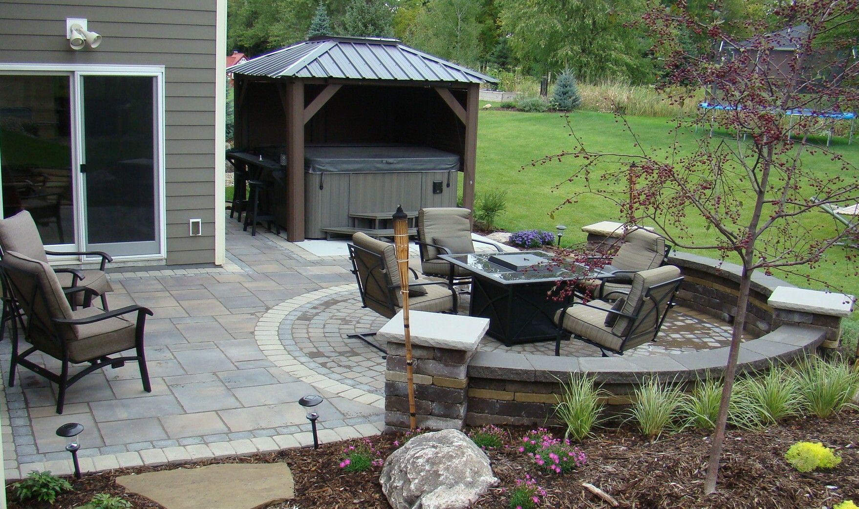 Patio with seating around a fire pit, hot tub under a gazebo, and house in the background.