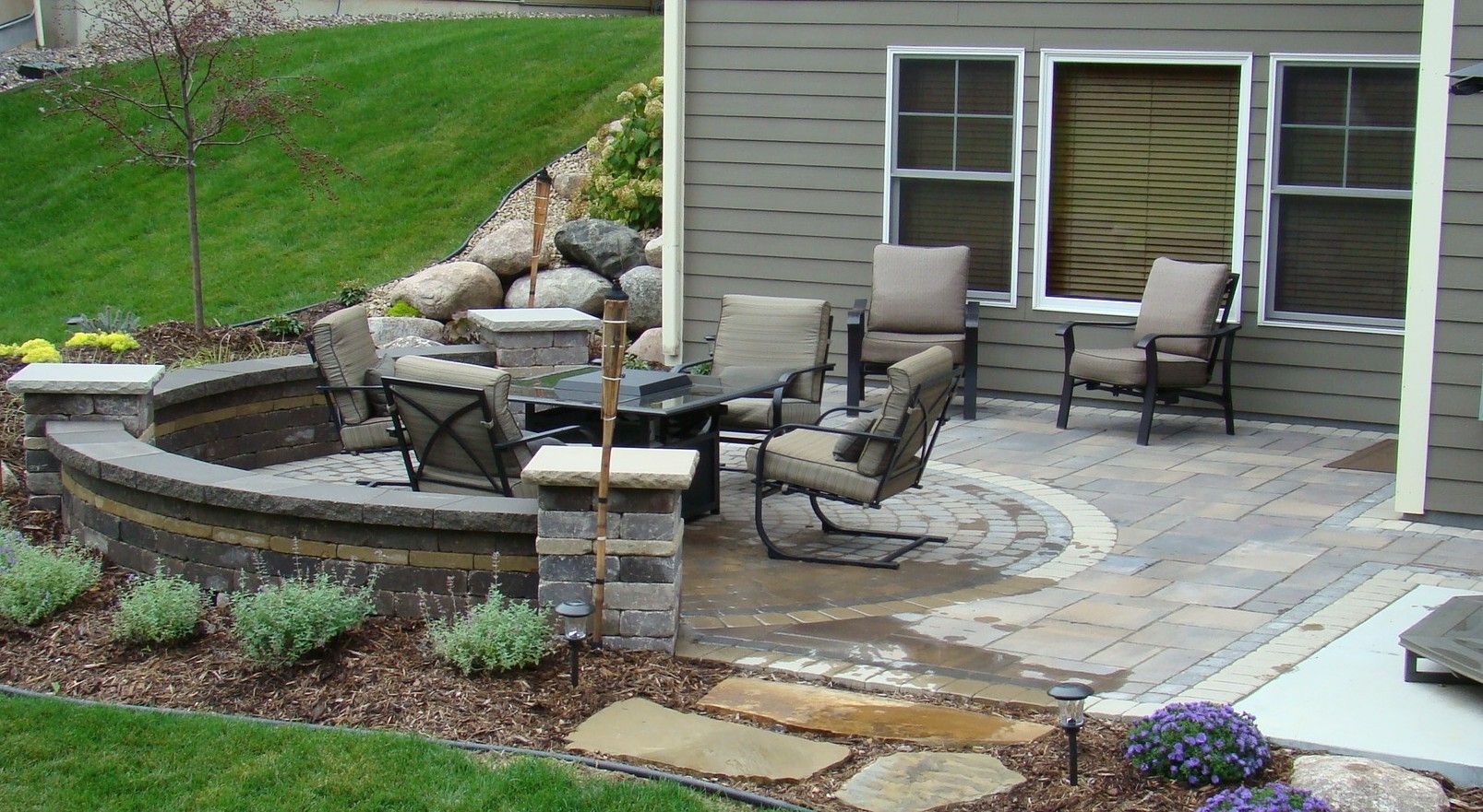 Outdoor patio with fire pit, seating, and stone pavers near a house.