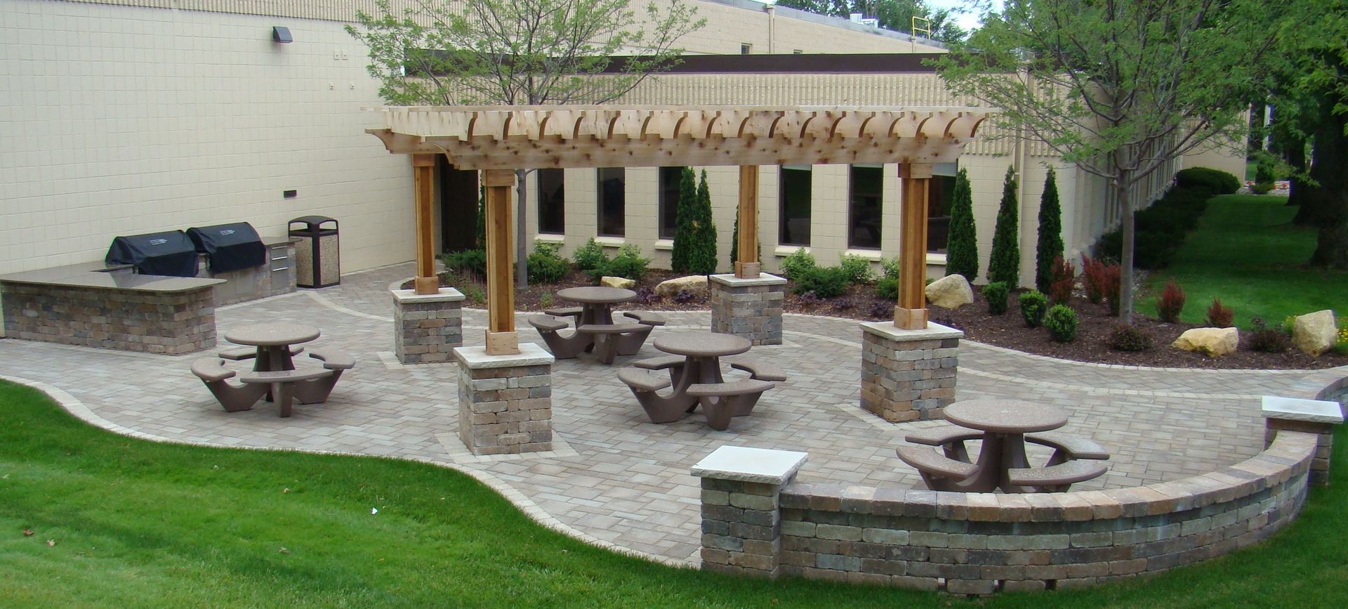Outdoor dining area with tables, pergola, and brick patio. Adjacent to green grass and a building.