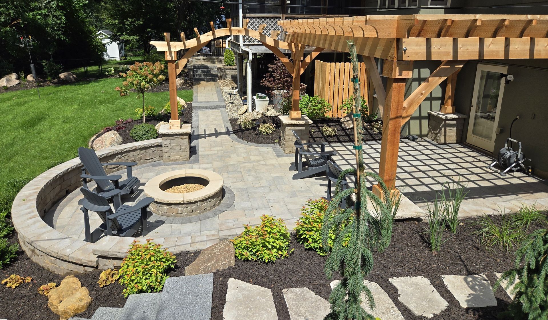 Outdoor patio with fire pit, seating, and wooden pergola. Paved area surrounded by landscaping.