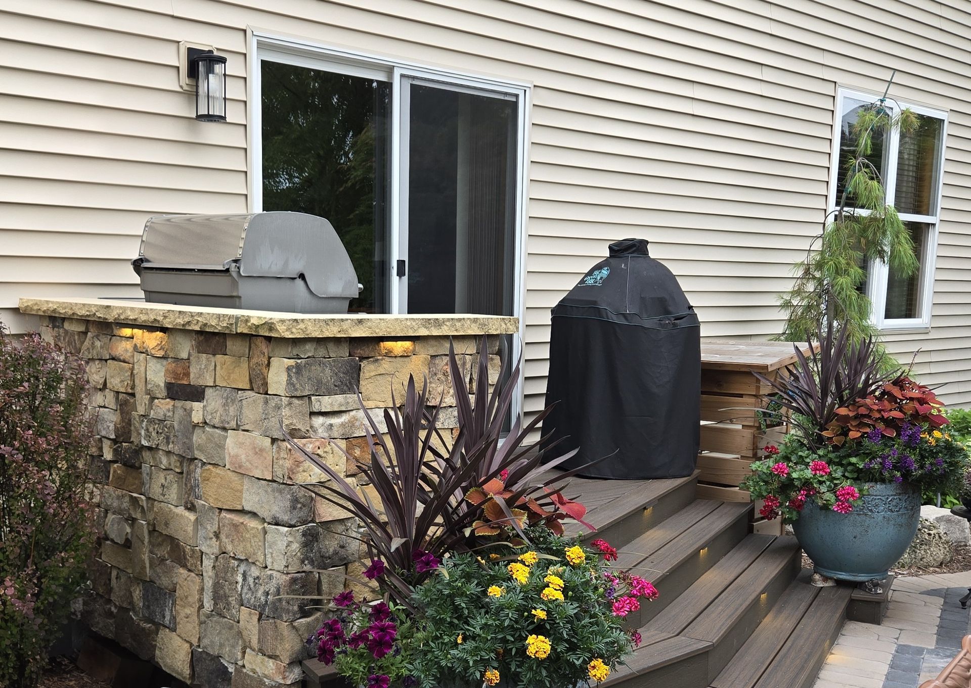 Stone outdoor kitchen with grill and smoker, near a house with deck and colorful flowers.