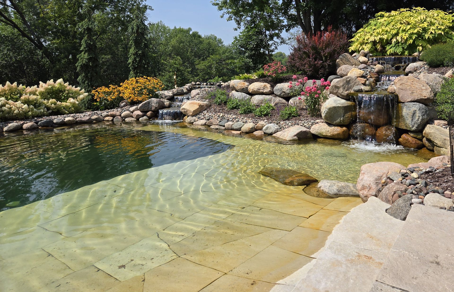 Pond with waterfall flowing over rocks; colorful flowers and plants surround the water.