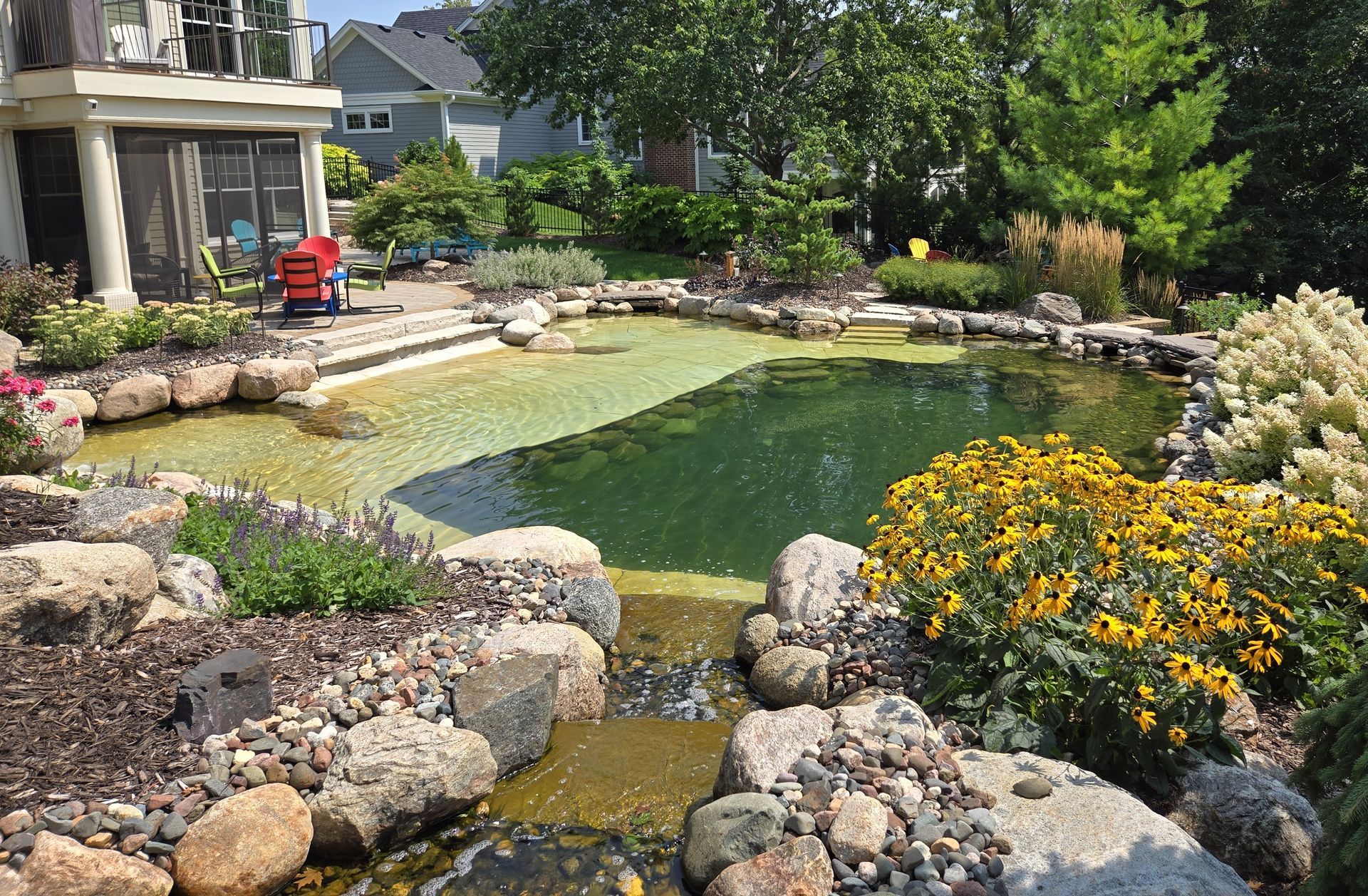 Stone-lined backyard pond with waterfall, surrounded by rocks and flowers. Lush green water and clear sky.