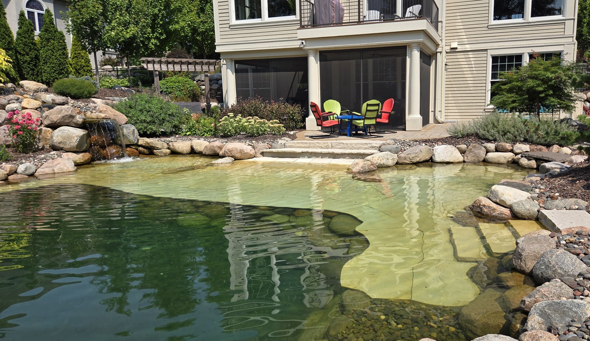 A pond with stone steps leads to a patio with colorful chairs in front of a house.