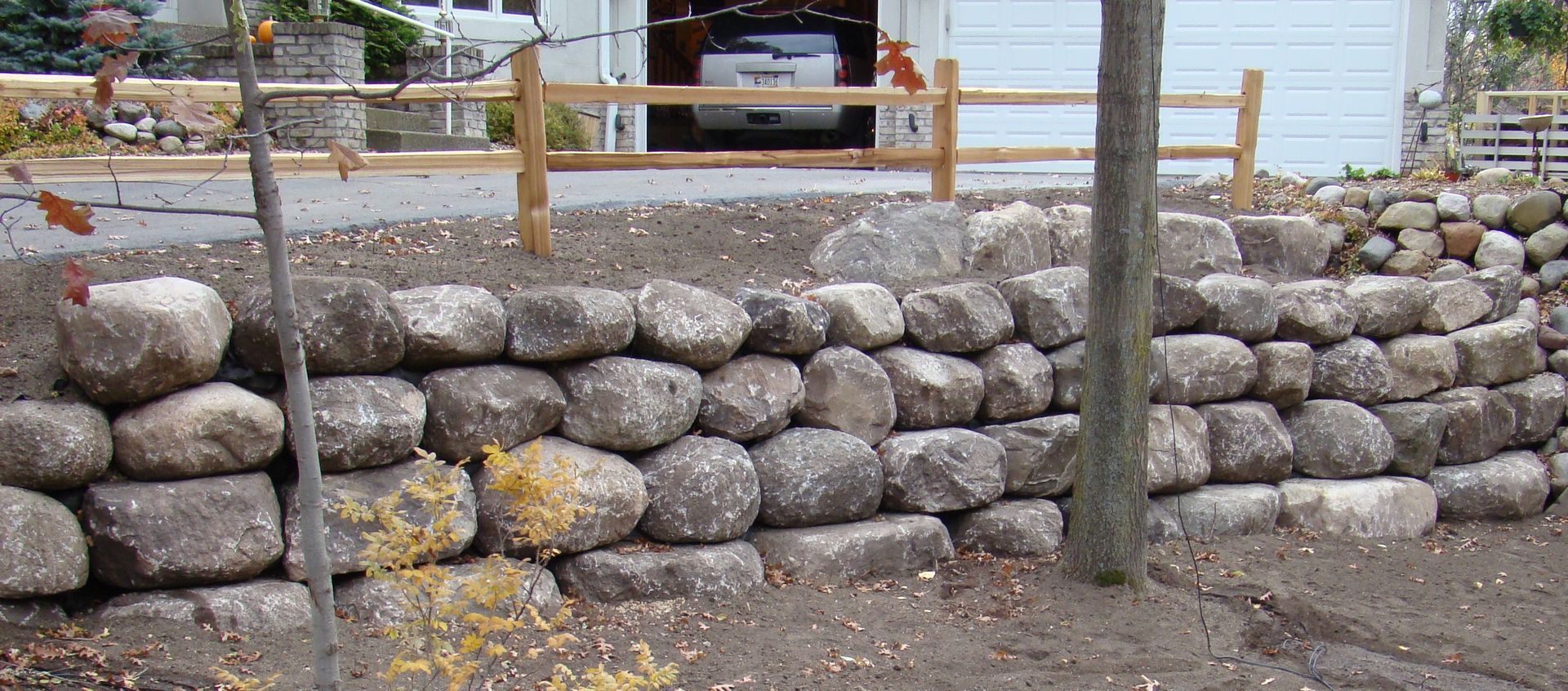A retaining wall made of large stones in front of a house, with a wooden fence and tree.