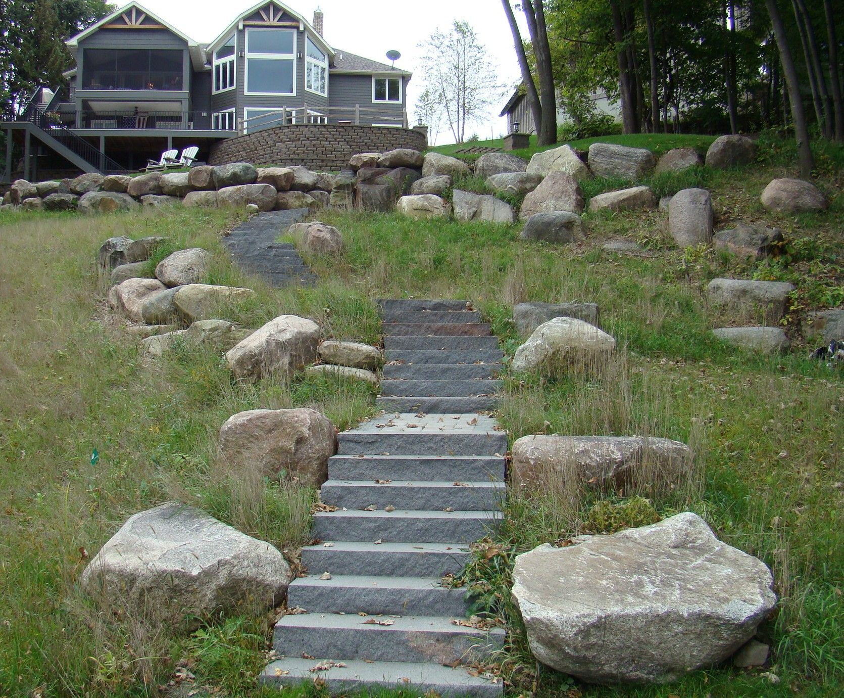 Stone steps leading uphill to a house with large windows, surrounded by rocks and grassy terrain.