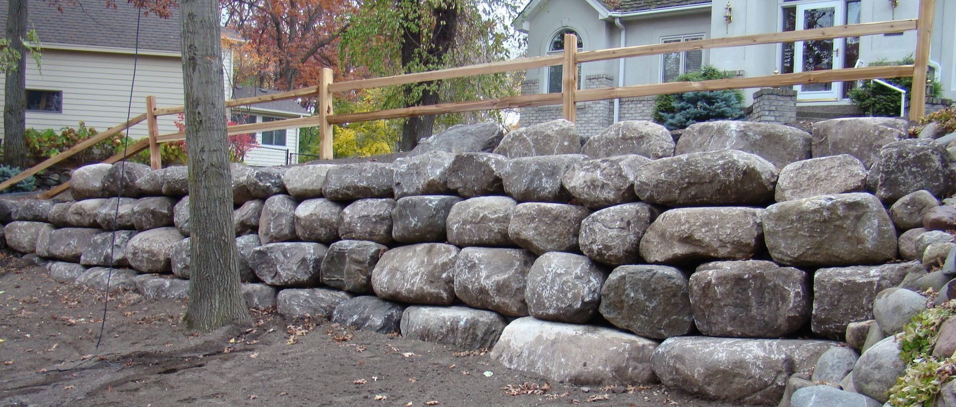 Stone retaining wall with a wooden fence in the background, near a house and trees.