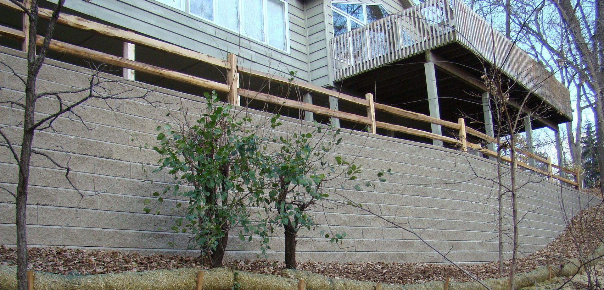 A multi-level gray retaining wall supports a house with a wooden deck and a split-rail fence.