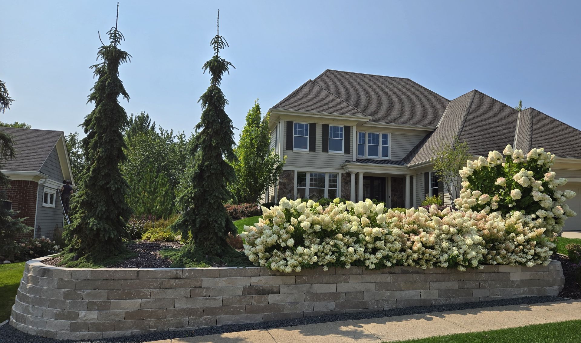 Two-story beige house with a stone retaining wall and ornamental trees and flowering bushes.