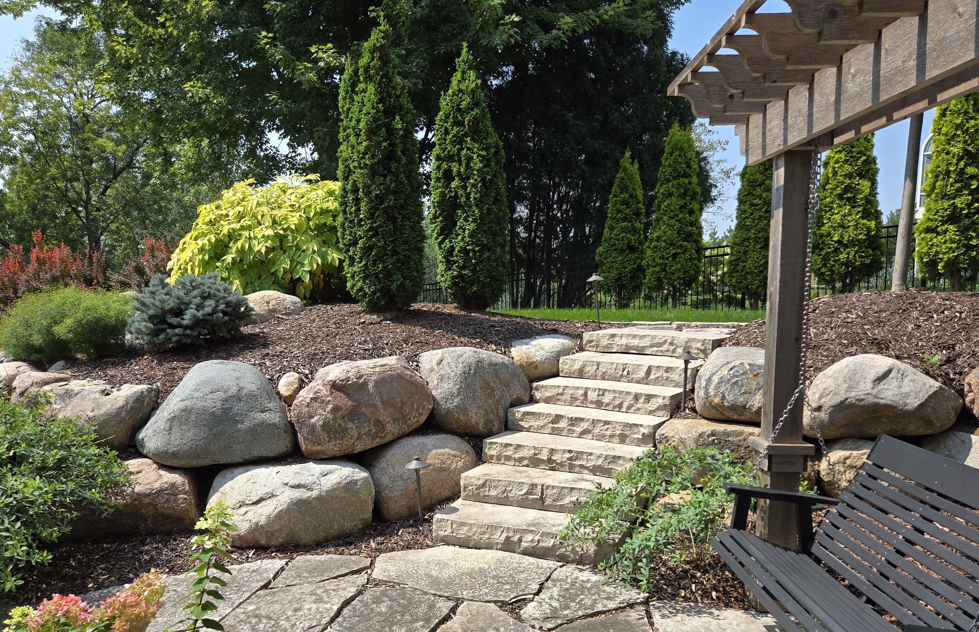 Stone steps ascend a terraced garden with large boulders, trees, and a wooden pergola on the right.