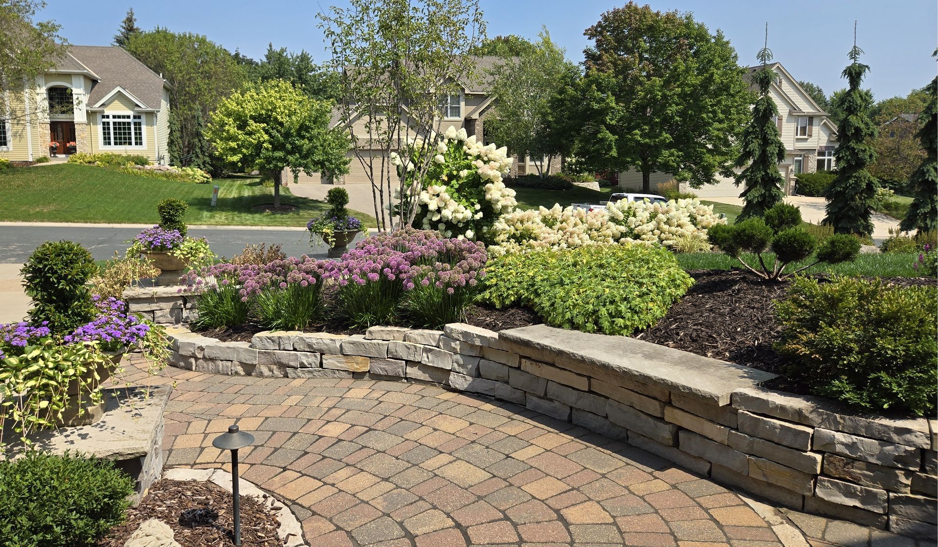 Brick pathway and stone wall with colorful flowerbeds, leading to houses on a sunny day.