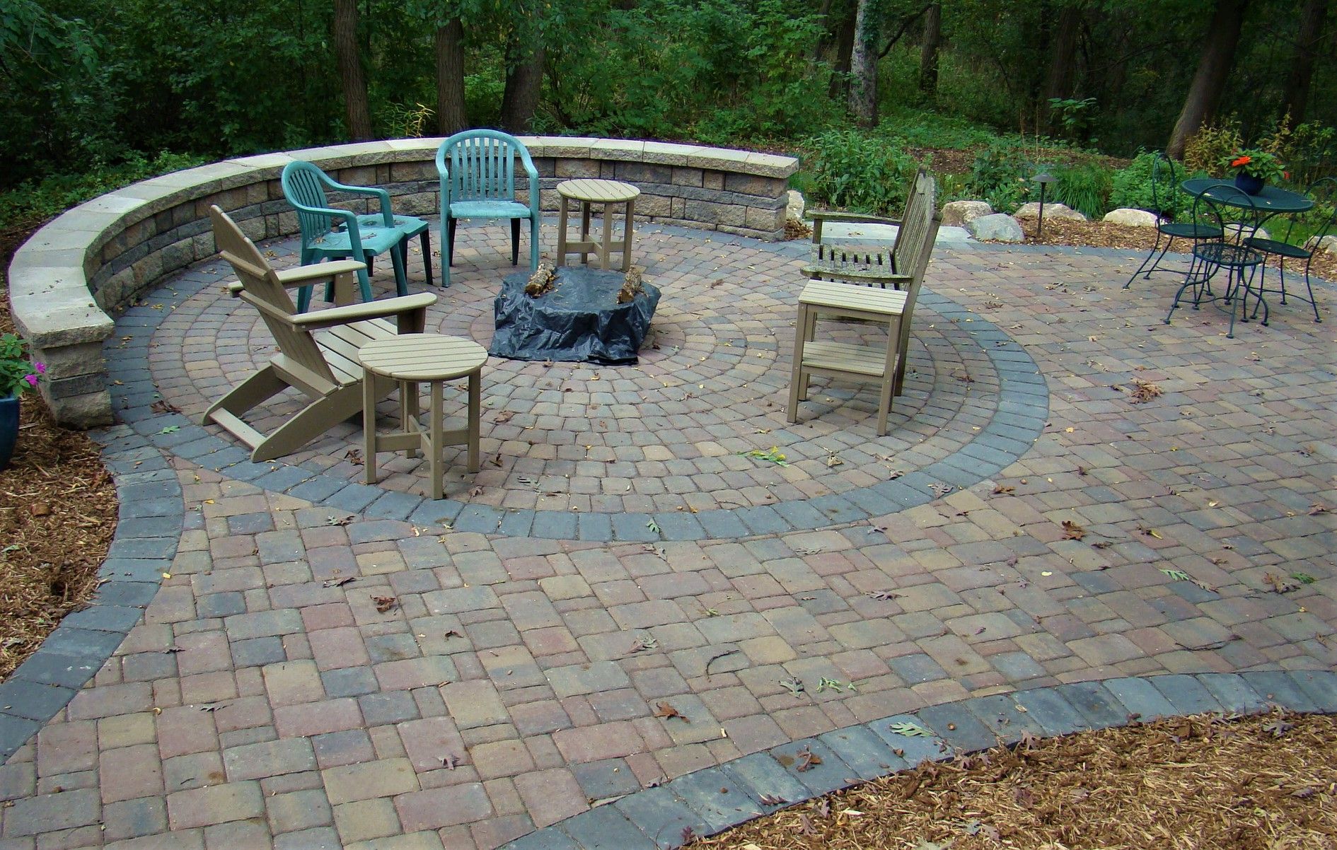 Brick patio with stone wall, chairs, and fire pit in a wooded backyard.