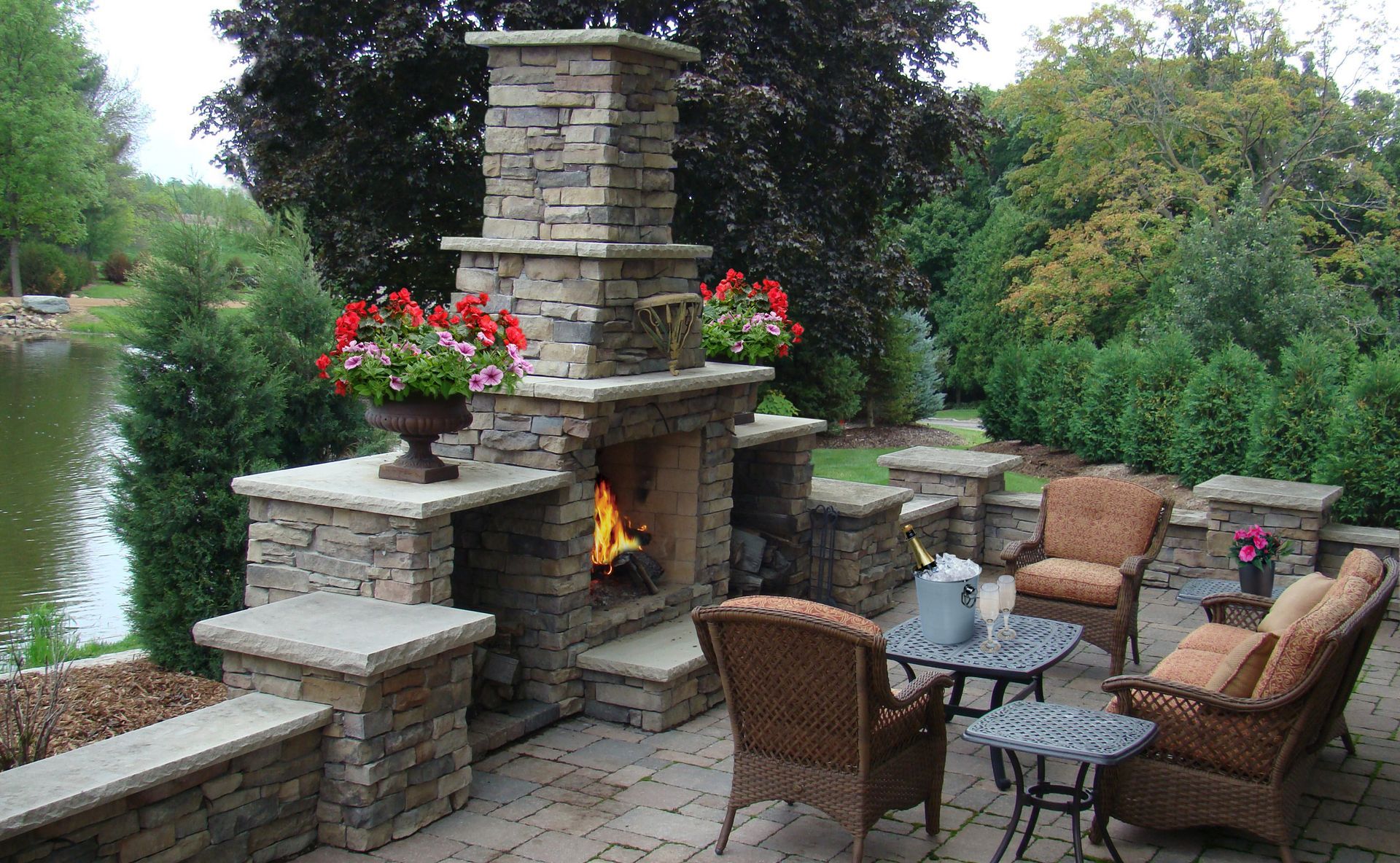 Stone fireplace with fire in a patio setting with seating and flowers, by a body of water.
