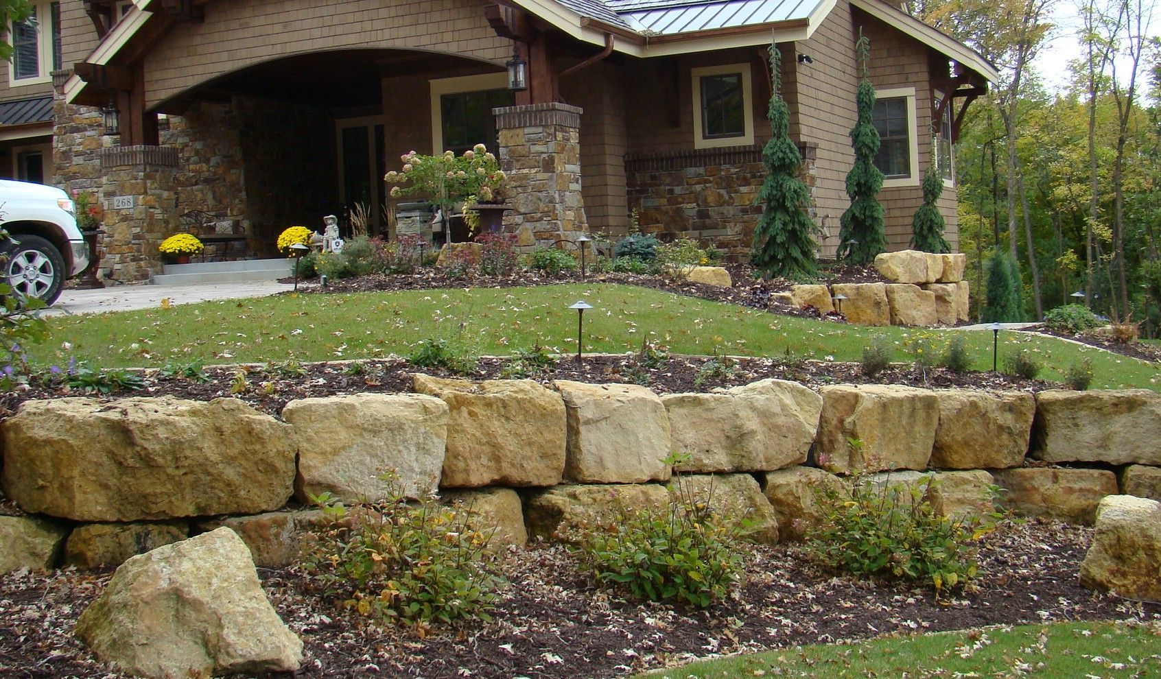 Stone retaining wall in front of a house, with a grassy yard and landscaping.