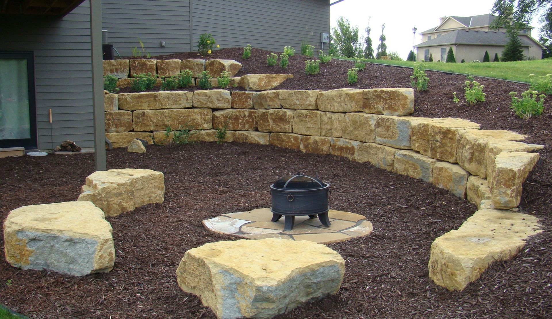 A fire pit area with stone retaining walls and seating, surrounded by mulch. A house is in the background.