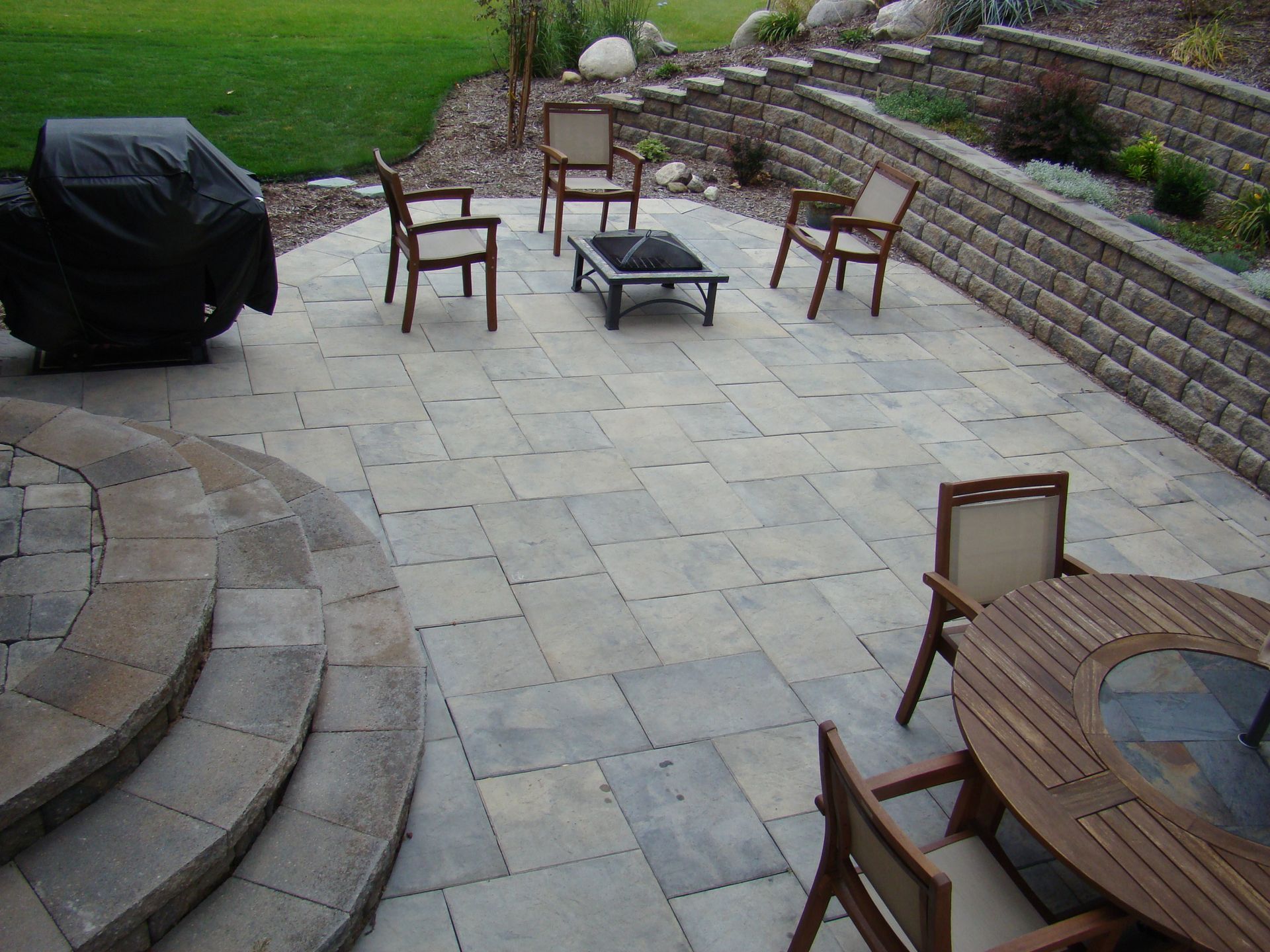 Patio with stone pavers, seating, fire pit, and grill near a retaining wall and grass.