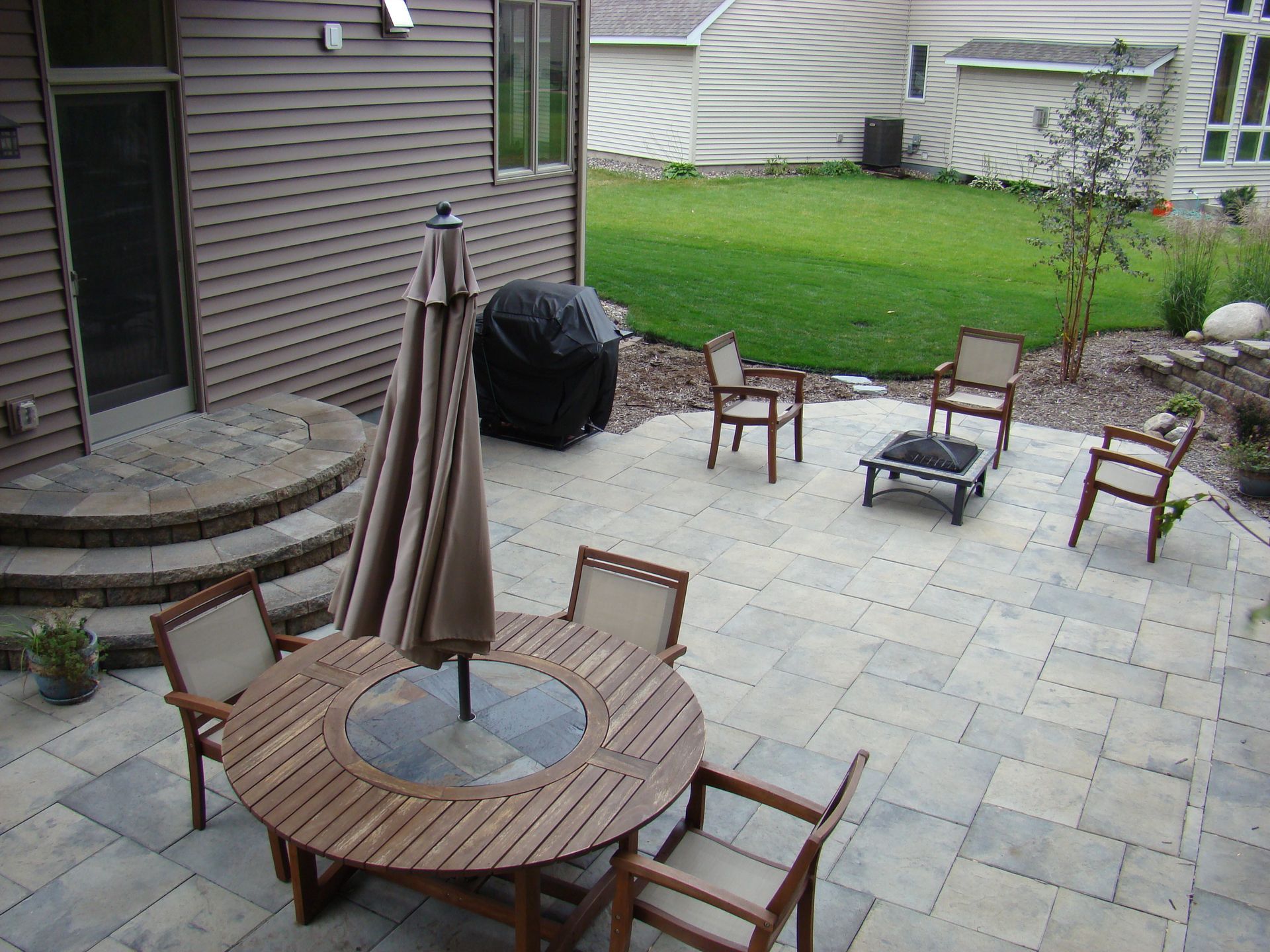 Patio with round wooden table, chairs, grill, umbrella, and stone pavers in a backyard setting.