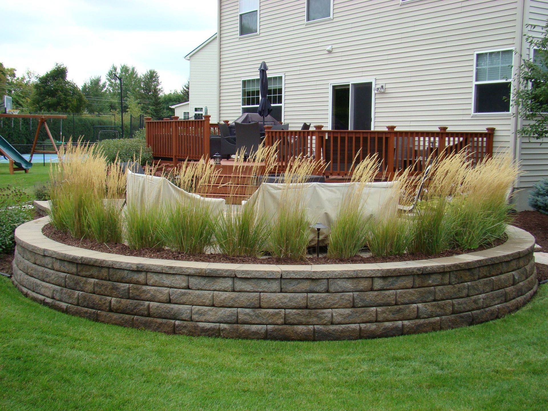 Curved retaining wall with grasses in front of a house's wooden deck. Green lawn surrounds.