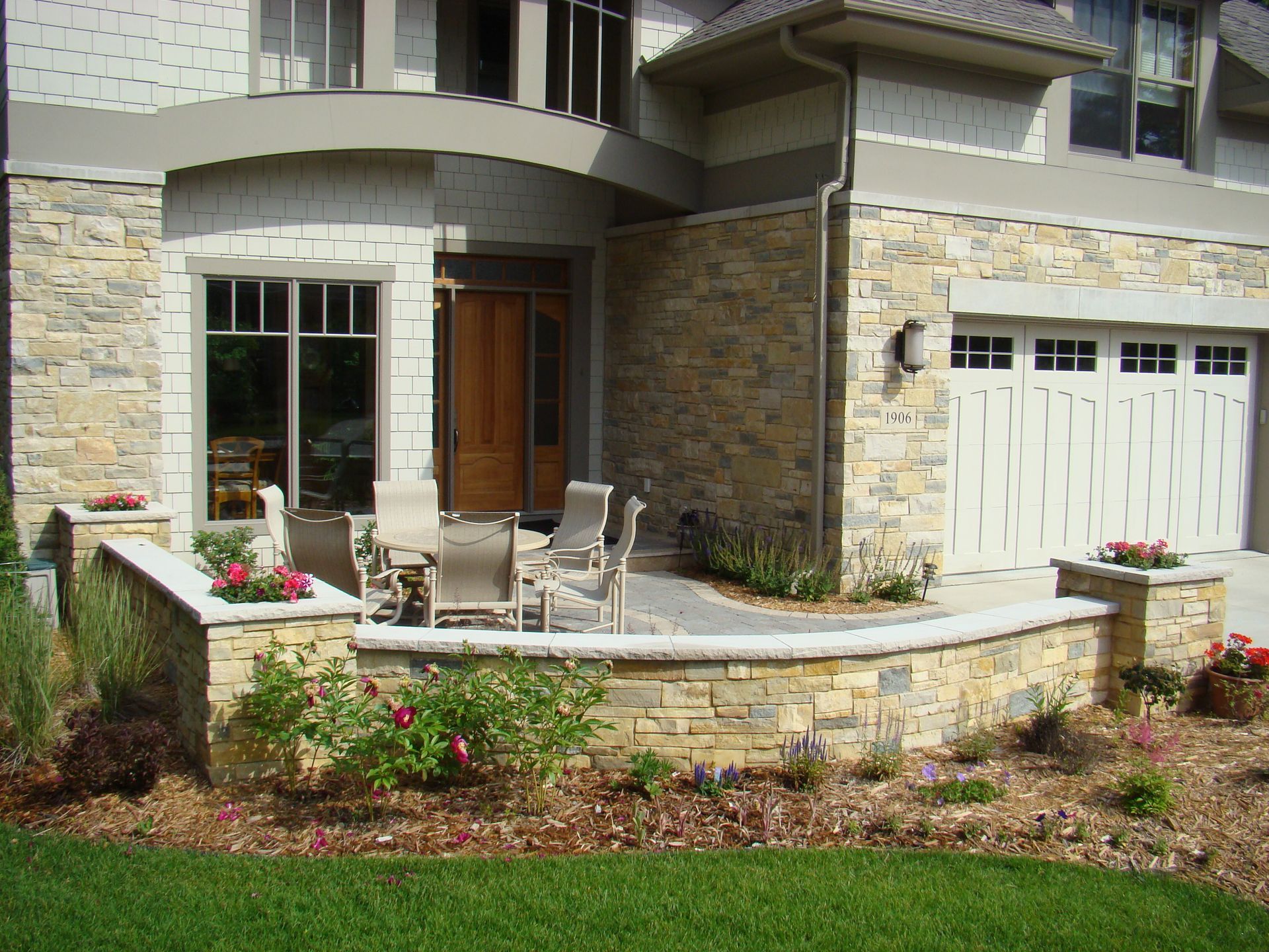 Stone exterior of a house with an arched entry, patio furniture, and flower beds.