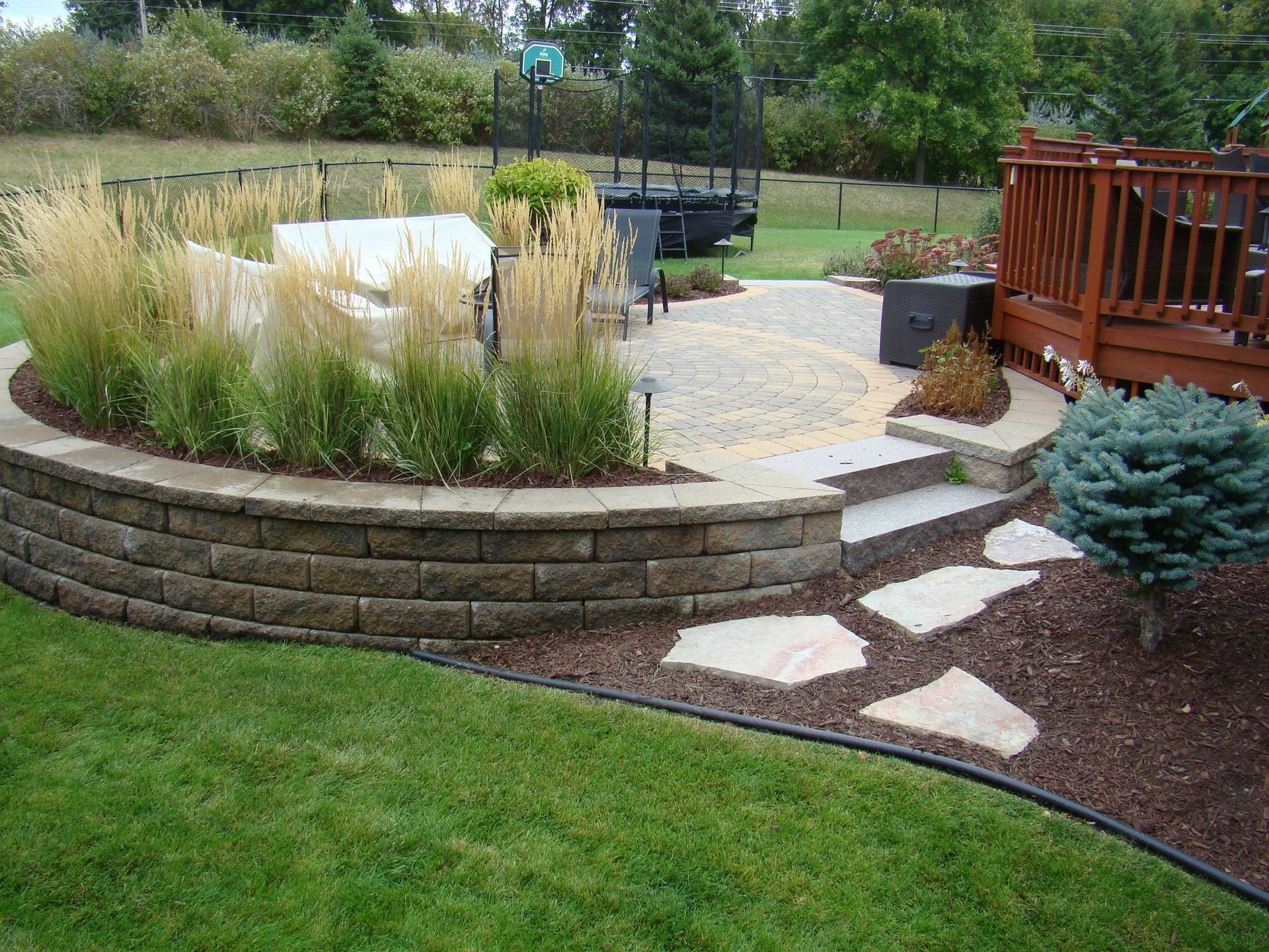 Curved retaining wall with patio, steps, and deck in a landscaped backyard.