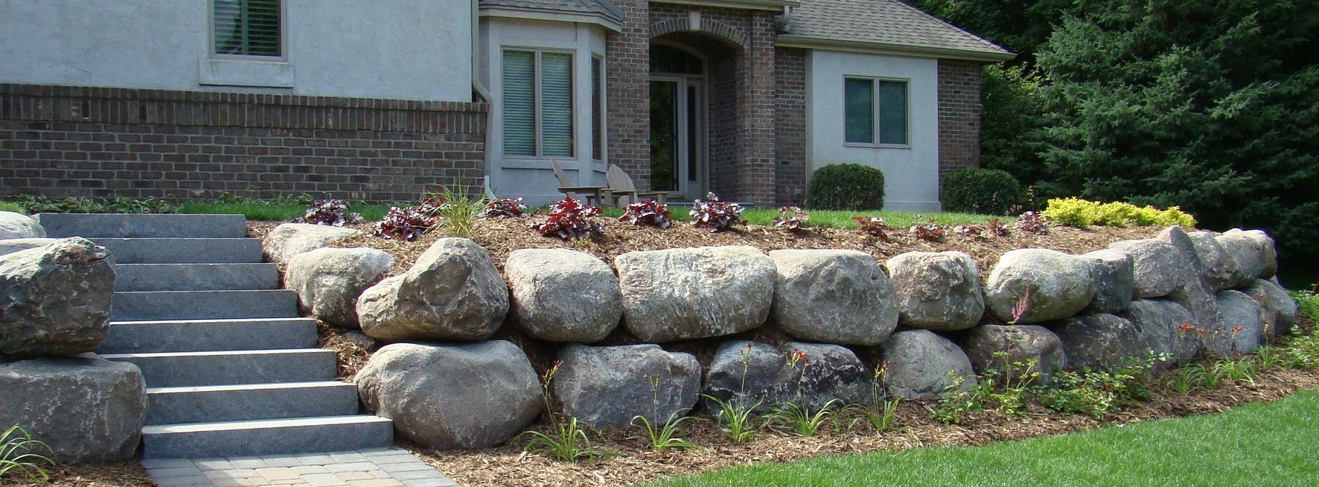 Stone steps leading up to a house with a rock wall and flower beds.