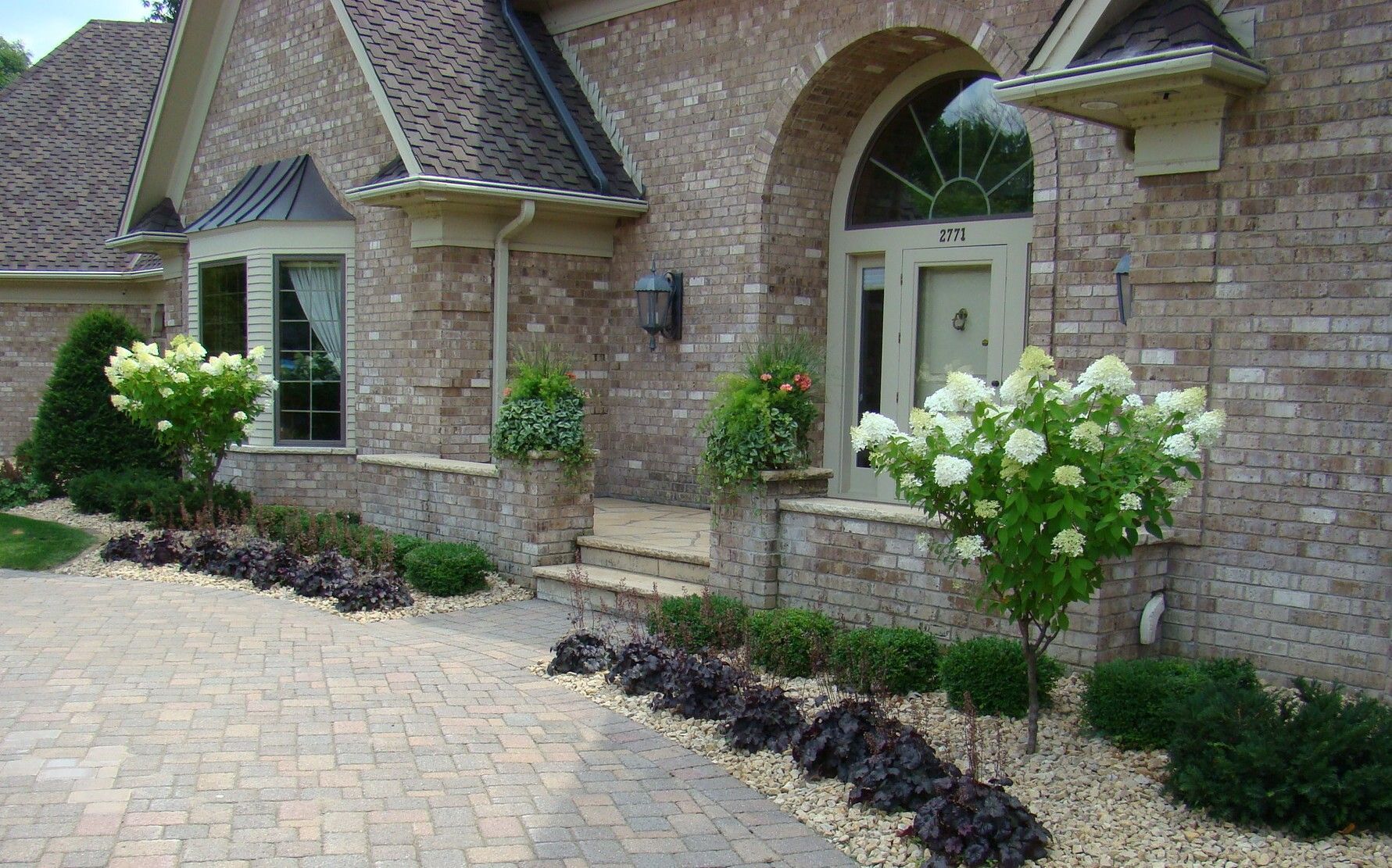 Brick home exterior with arched doorway, stone path, landscaping with flowering bushes and dark mulch.