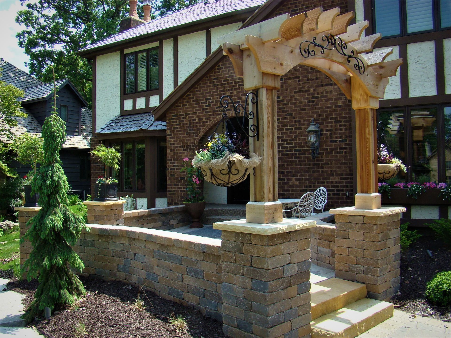 A brick home entrance with an arched trellis, stone walls, and a hanging flower basket.