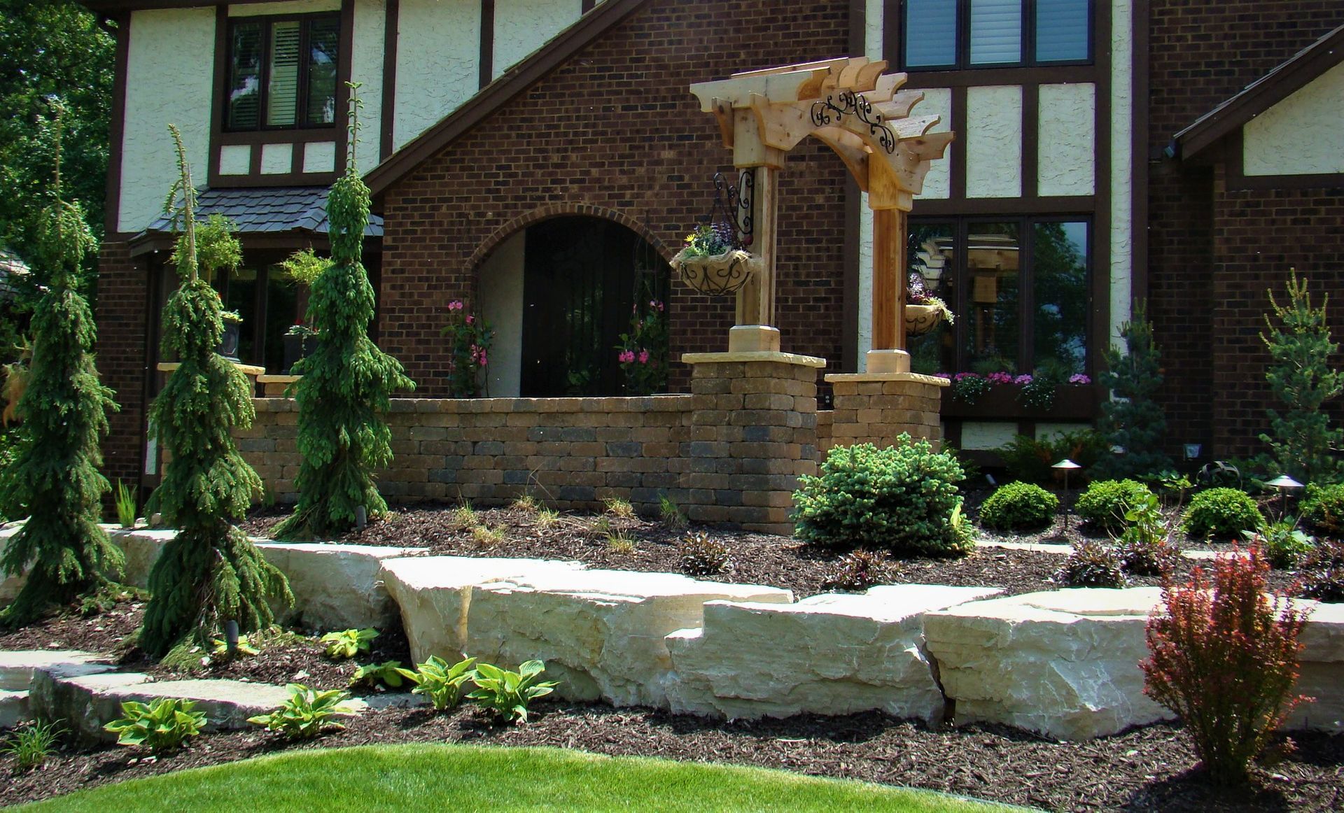 Stone retaining wall in front of a brick house with a pergola and landscaping.