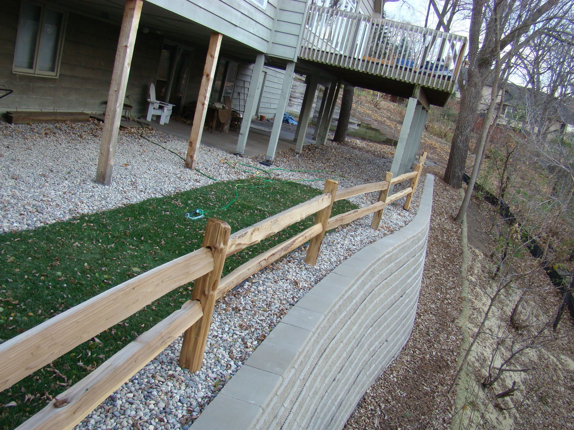 Wooden split-rail fence alongside a concrete retaining wall, separating a grassy area from a wooded hillside, near a deck.
