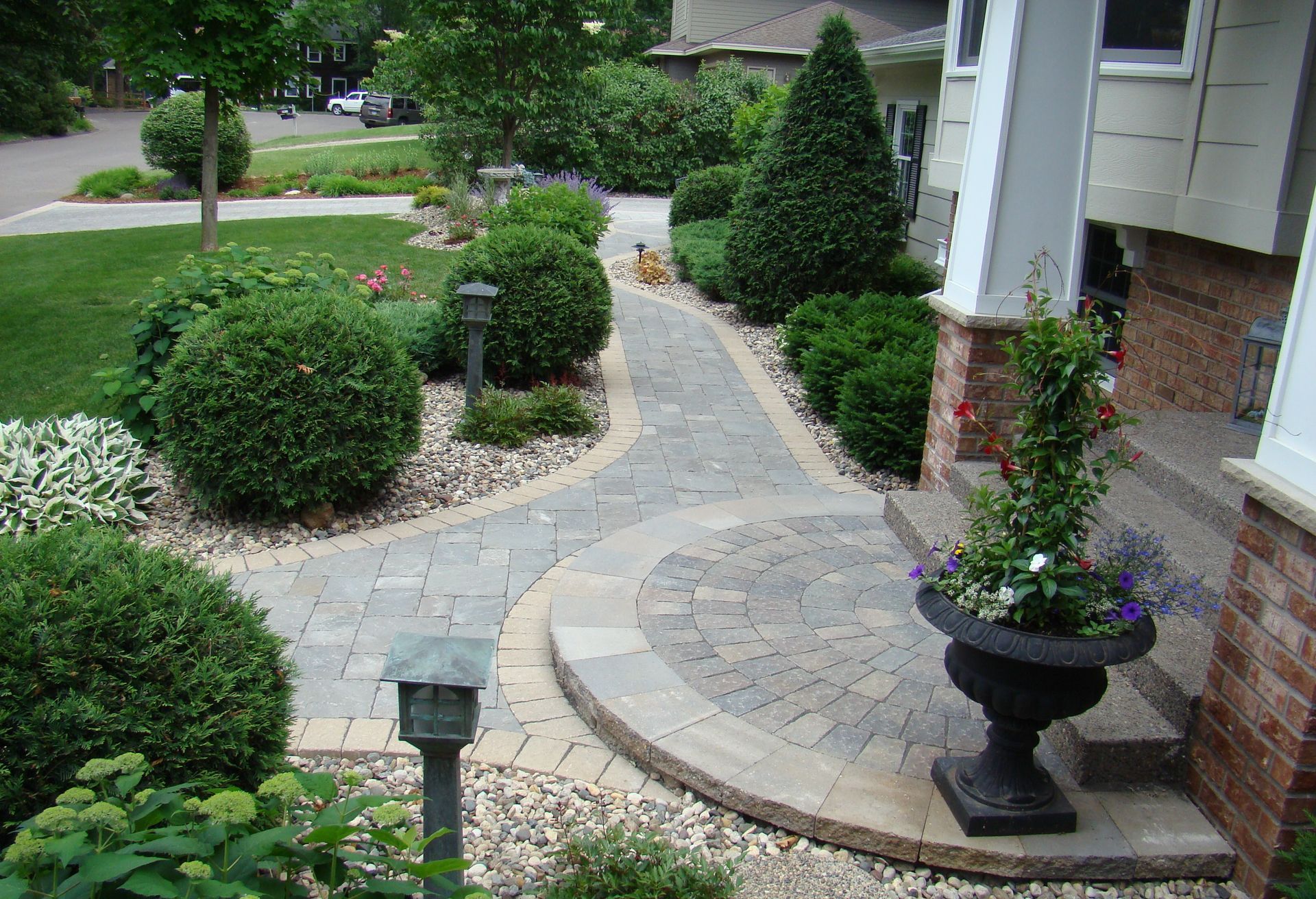Paver walkway curving towards a house, lined with green shrubs and decorative potted flowers.