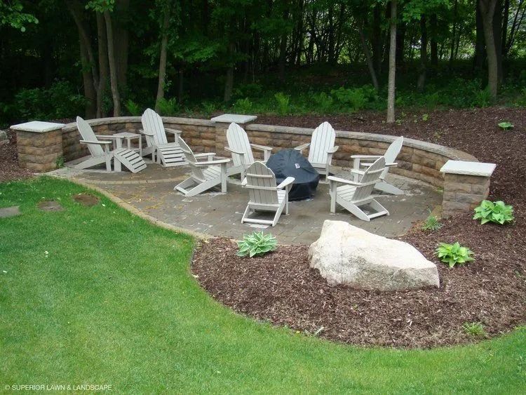 Outdoor patio with chairs around a fire pit, surrounded by a low stone wall, in a yard with trees.