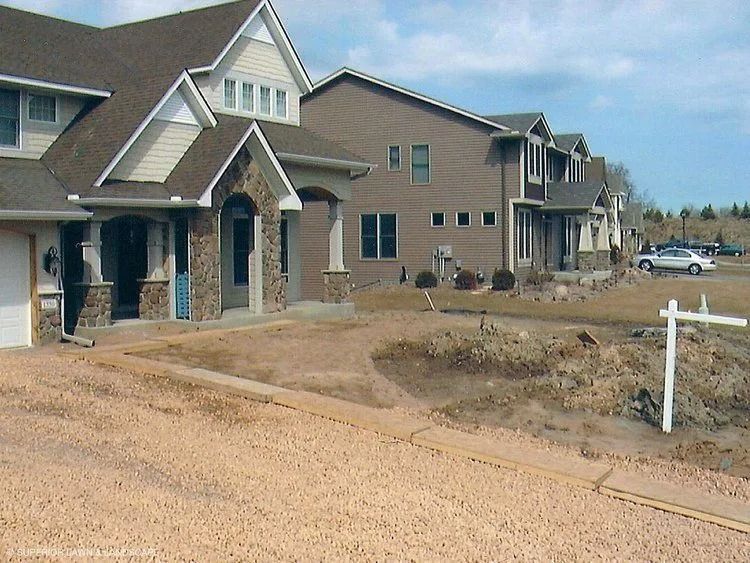 Houses under construction; beige siding, stone accents, gravel driveway, and a white sign.