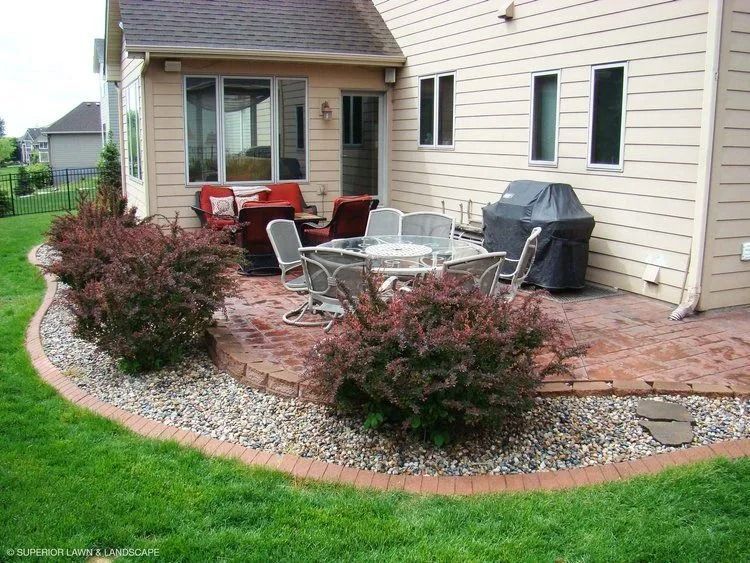 Patio with a brick border, red stone, shrubs, and outdoor furniture next to a tan house.