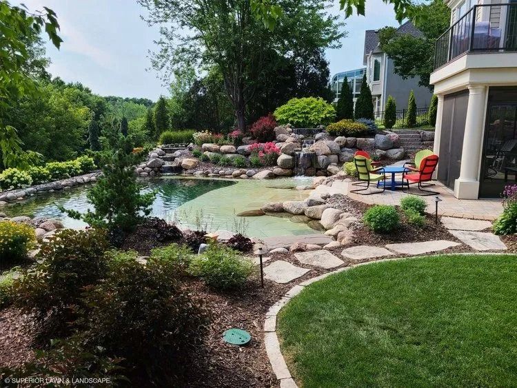 Landscaped backyard with pond, stone pathway, colorful flowers, and patio furniture.