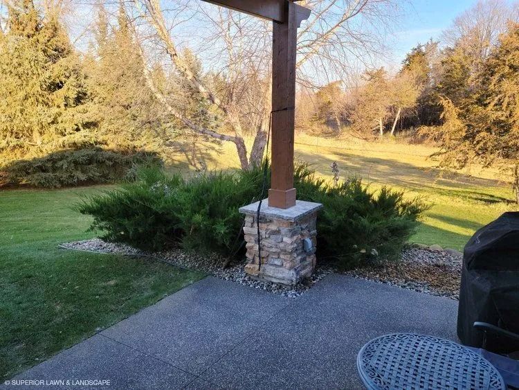 A backyard patio with stone column, green bushes, and trees in the background.