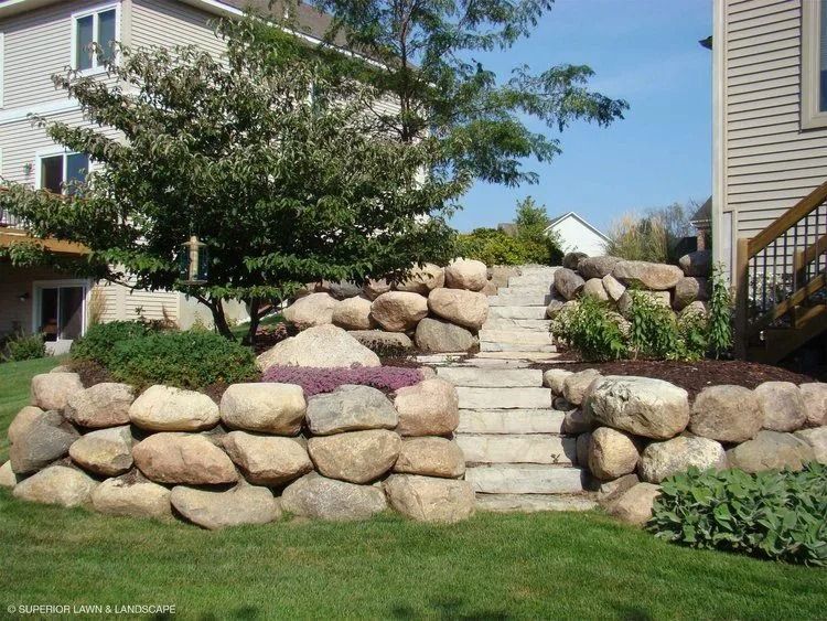 Stone retaining wall with steps leading uphill to a house. Landscaped with plants and a tree.