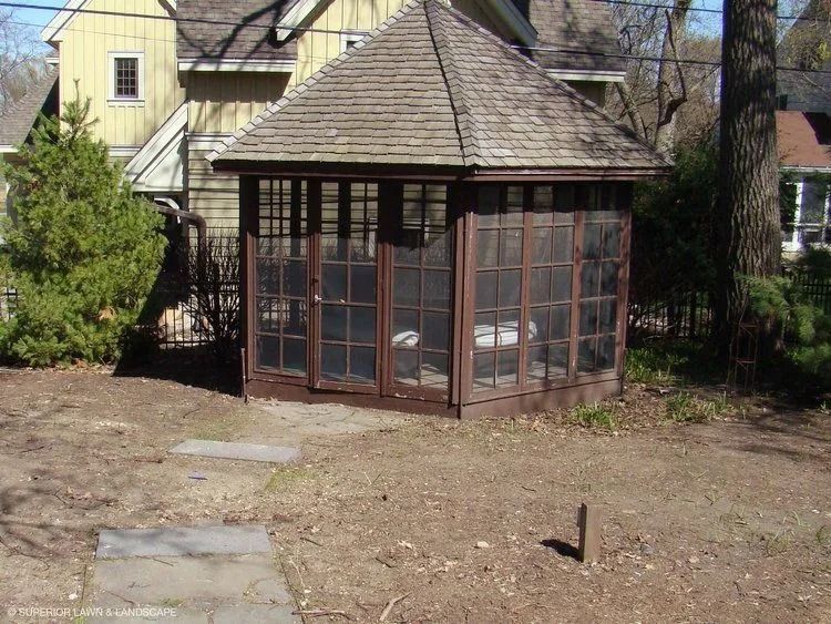 Brown wooden gazebo with glass windows and a shingled roof, in a yard.