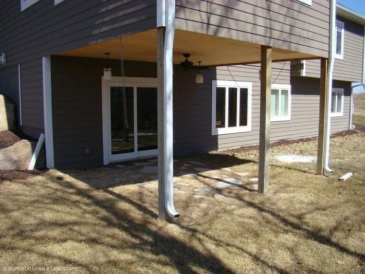 Under deck patio with glass sliding door and two windows. Brown siding and grass with two support beams and gutter.