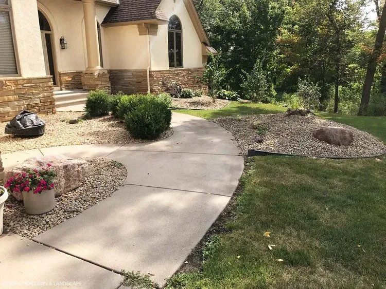 Curving concrete walkway leading to a house with landscaping, including bushes and rock beds.
