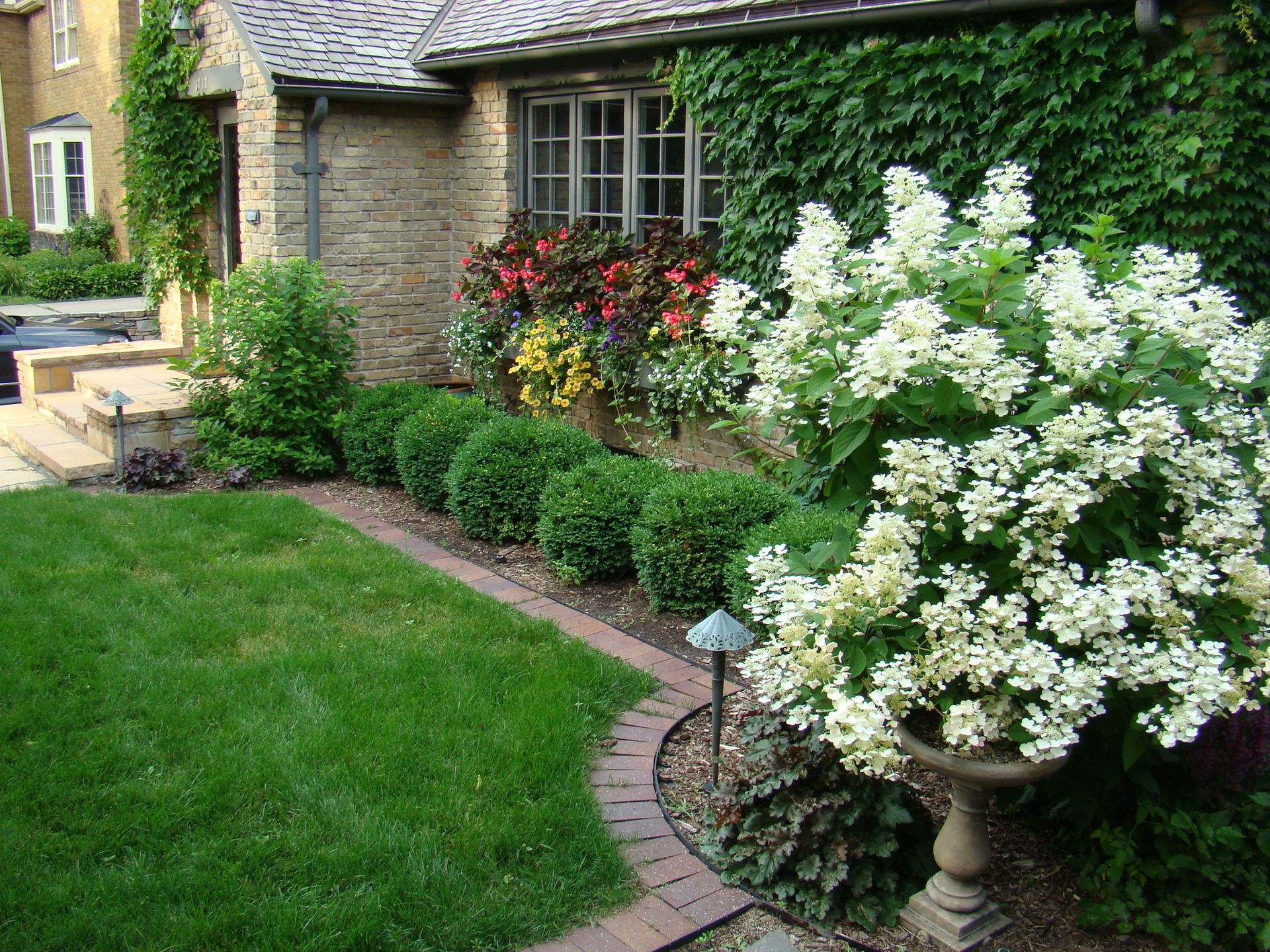 A house exterior with green lawn, flowerbeds, and a large white flowering plant in a decorative urn.