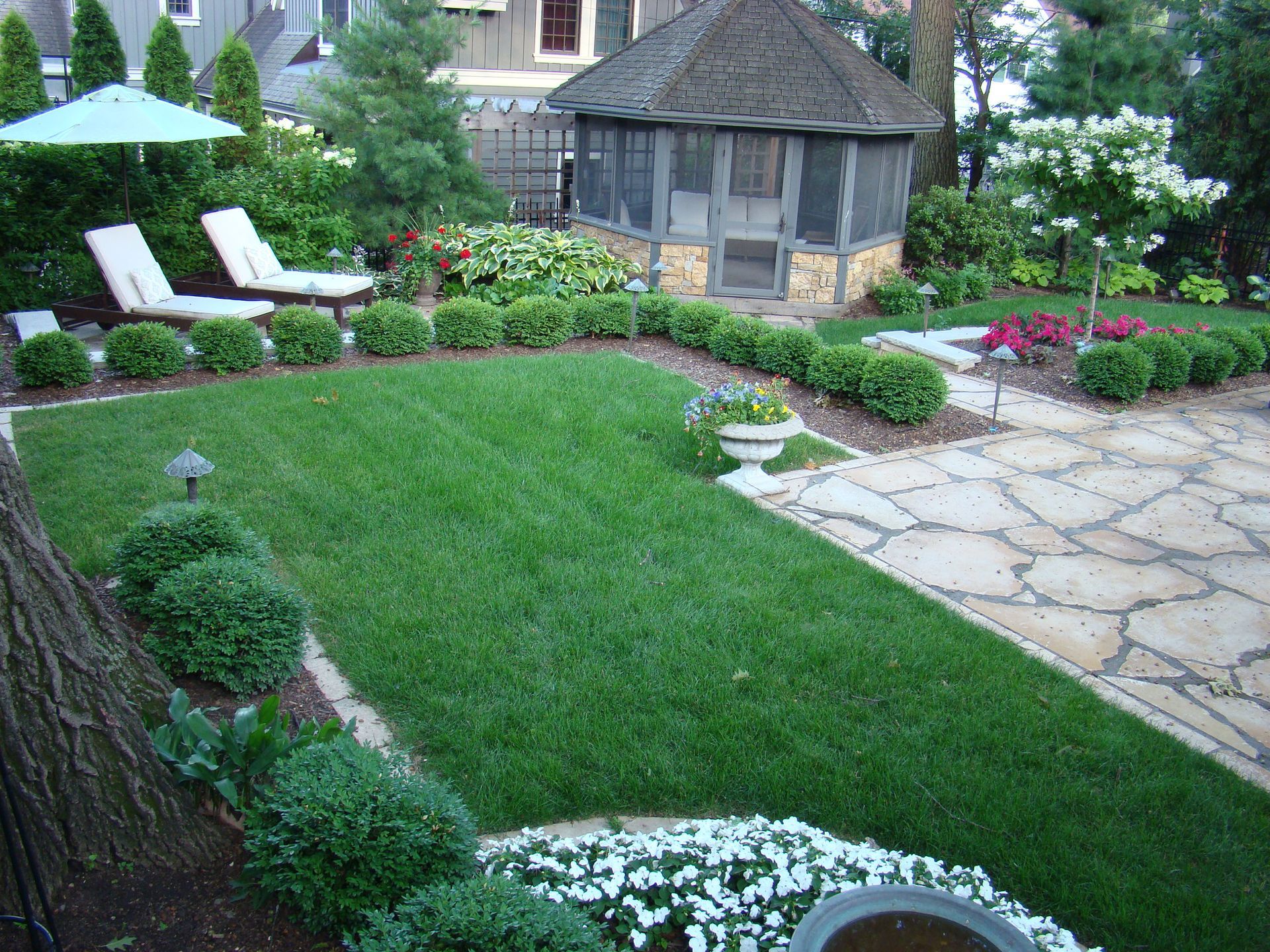 Lawn with manicured shrubs, a stone patio, gazebo, lounge chairs, and garden.