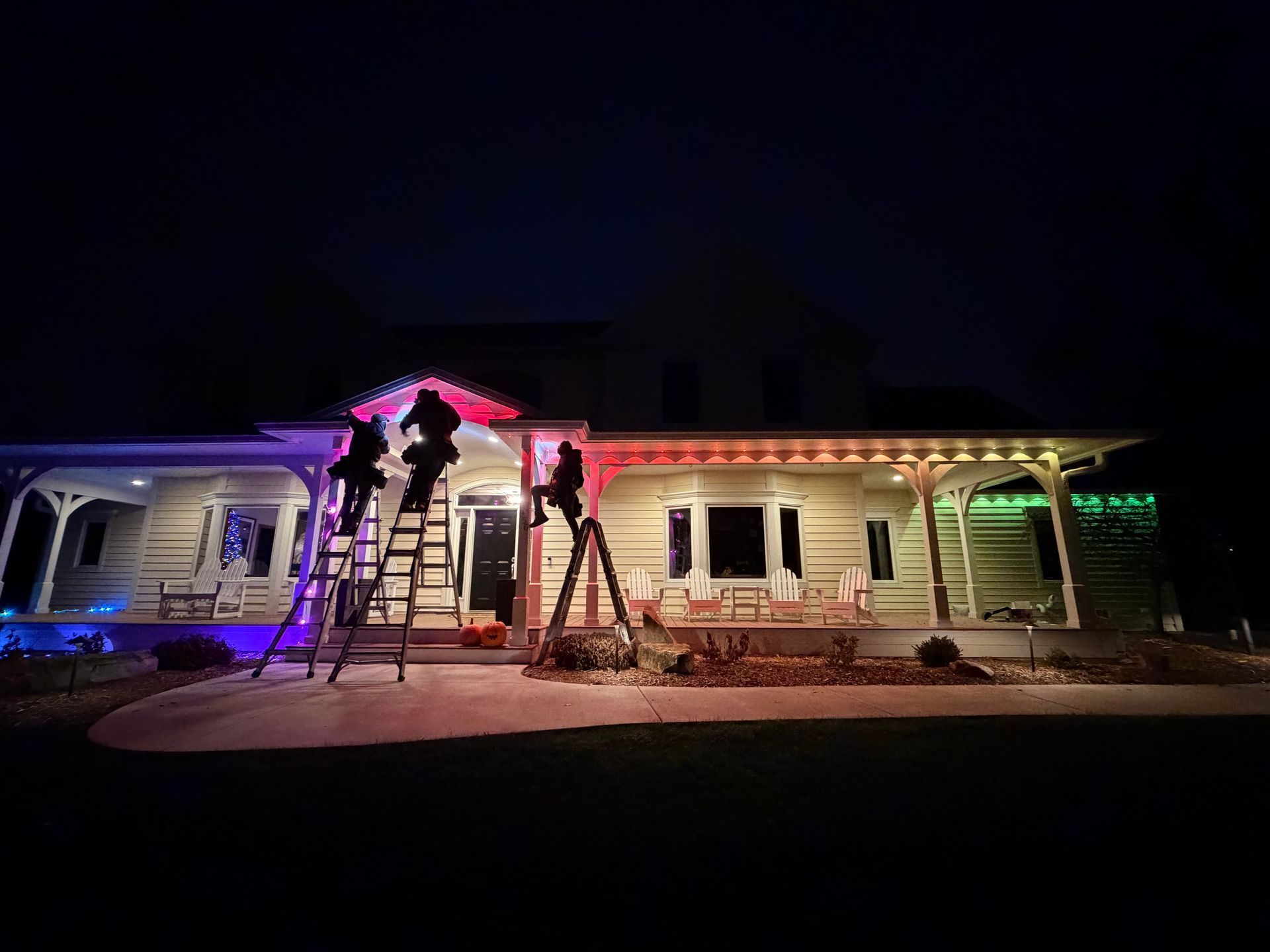 People on ladders are installing colorful lights on a house's roof at night.