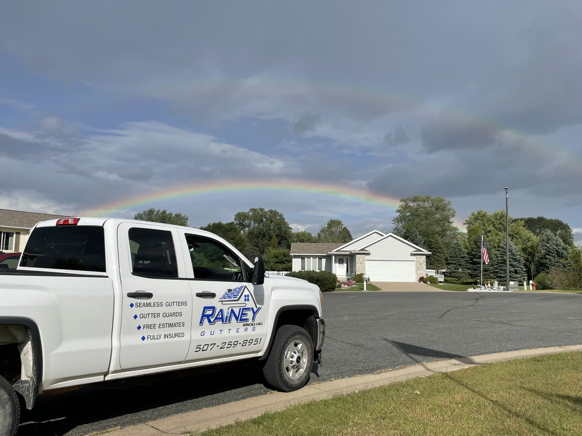 White work truck parked near a house with a rainbow arcing in the sky.