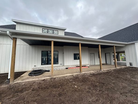 Exterior of a house under construction with a covered patio. White siding, black roof, wooden support beams, overcast sky.
