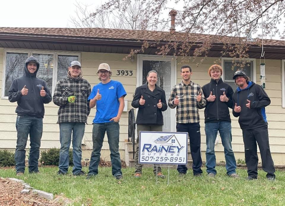 Group of people in front of a house, thumbs up. Rainey sign in front. Cloudy day.