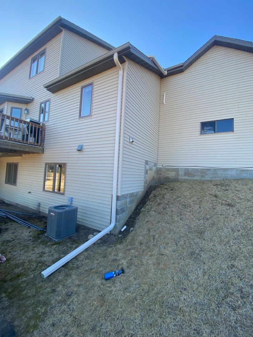 Beige house exterior with a white gutter system and a grassy hill on a sunny day.