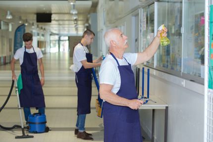 Three people wearing blue aprons cleaning a hallway; one vacuums, one wipes a window, and the other sprays a window.