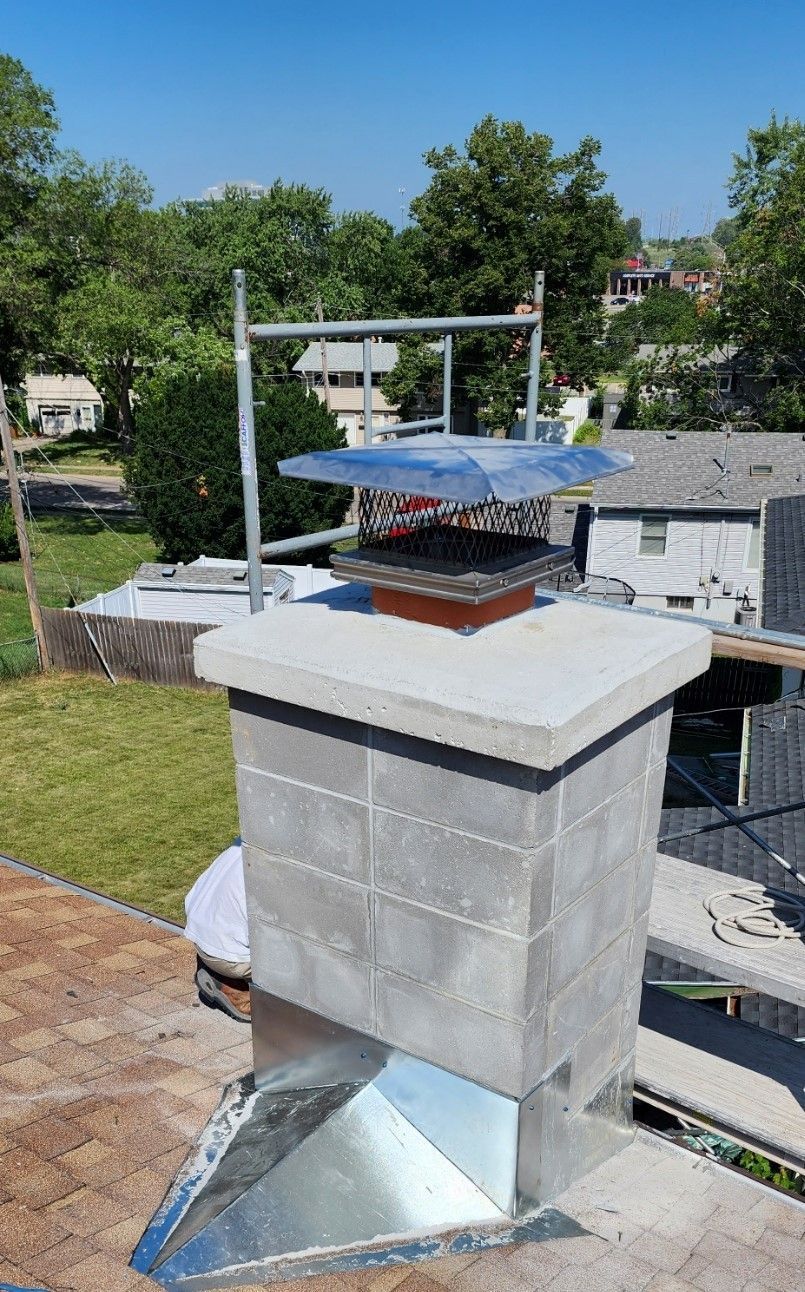 Concrete chimney with cap and roof flashing against a green tree backdrop.