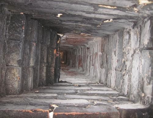 Inside view of a dark, rectangular brick chimney. Walls covered in soot, leading to a distant light.