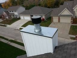 White-sided chimney with silver cap and black top on a rooftop, suburban houses in the background.
